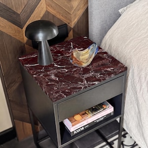 May include: A black bedside table with a dark red marble top, featuring a single drawer and an open shelf below. On the surface sits a black mushroom-shaped lamp and a small, clear glass bowl with blue and amber swirls. Two books are on the shelf.