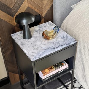 May include: A black bedside table with a white marble top, featuring a matte black mushroom-shaped lamp and a small, curved, multi-coloured glass bowl. The table has a single drawer and an open shelf containing two books titled Icons and Big Bag.