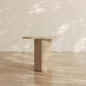May include: A beige stone table featuring a smooth, circular tabletop supported by a central pedestal base. The pedestal is constructed with vertical, fluted panels that create a textured, column-like appearance throughout the entire structure.