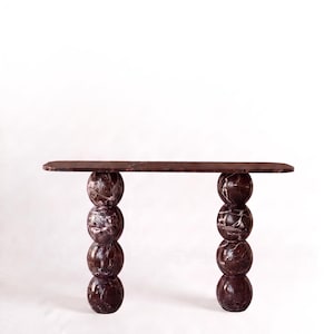 Op de afbeelding: Een consoletafel gemaakt van donker roodbruin marmer met witte aders. De tafel heeft een lang, rechthoekig blad dat wordt ondersteund door twee verticale kolommen, elk bestaande uit vier gestapelde marmeren bollen van dezelfde steen.