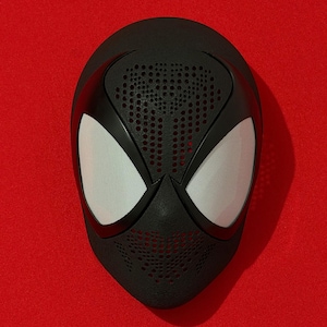 Peut inclure: Un masque intégral noir doté de grands verres en maille blanche en forme d'amande. La surface du masque présente une texture composée d'une densité de petites perforations circulaires sur le front et le menton, créant un aspect pointillé.