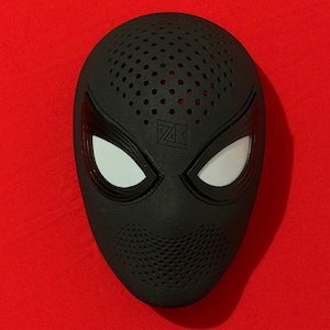 May include: A black full-face mask featuring a textured surface with numerous small ventilation holes across the forehead and chin areas. The mask includes two large, almond-shaped eye openings covered with white mesh lenses and a logo.