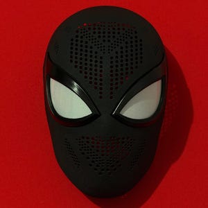 May include: A matte black full-face mask featuring a textured surface with numerous small circular perforations across the forehead and mouth areas. The mask includes two large, almond-shaped eye openings covered with a fine white mesh material.