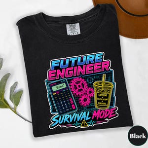 May include: A black t-shirt featuring a graphic print with the text "FUTURE ENGINEER SURVIVAL MODE". The design includes a calculator displaying "E=mc2", three pink gears, and a drink cup labeled "CAFFEINE + CALCULATIONS = SURVIVAL".
