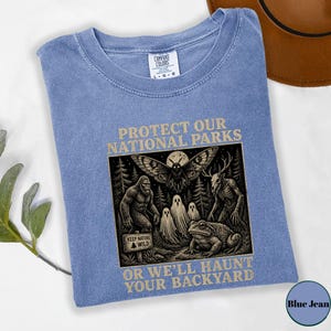 May include: A blue crew neck t-shirt featuring a graphic print with the text PROTECT OUR NATIONAL PARKS and OR WE'LL HAUNT YOUR BACKYARD. The illustration depicts a moth, a sasquatch, ghosts, a deer-like creature, and a toad in a forest.