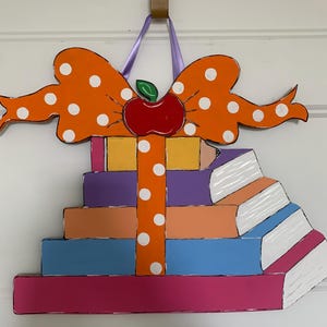 May include: A decorative wooden wall hanging shaped like a stack of five colorful books in pink, blue, peach, purple, and yellow. An orange ribbon with white polka dots wraps around the books, topped with a large orange bow and a red apple.