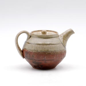 May include: A ceramic teapot featuring a two-tone glaze with a light beige upper section and a dark reddish-brown lower section. The body has a textured, ribbed surface pattern throughout, a curved handle, a short spout, and a matching lid.