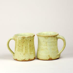May include: Two handmade ceramic mugs with a pale yellow and light green glaze featuring a speckled texture. Each mug has a sturdy handle and a slightly flared rim, showcasing a rustic, organic shape with visible variations in the clay finish.