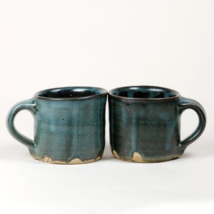 May include: Two ceramic mugs with a dark blue and teal glaze featuring a textured, dimpled surface. Each mug has a sturdy handle and a visible, unglazed light tan base. The glaze shows variations in color depth, creating a mottled appearance.