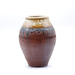 May include: A ceramic vase with a bulbous shape, featuring a textured reddish-brown body with a cross-hatched pattern. The upper section has a smooth, cream-colored glaze that drips down over the dark, textured surface of the vessel.