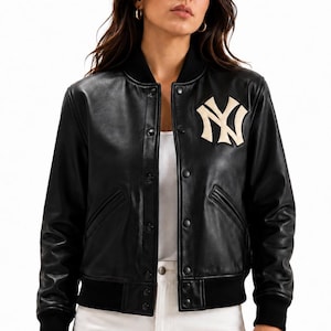 May include: A black leather varsity jacket featuring a white New York Yankees NY logo patch on the left chest. The jacket has a ribbed black collar, cuffs, and hem, with a front snap button closure and two side pockets over a white shirt.