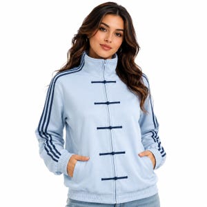 May include: A light blue zip-up jacket featuring a high collar, long sleeves with three dark blue stripes, and a front closure with five dark blue toggle-style frog fasteners. The jacket includes ribbed cuffs and a ribbed hem with two side pockets.