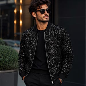 Può includere: Un giubbotto bomber nero ricoperto di piccole paillettes riflettenti che creano un effetto scintillante. La giacca presenta colletto, polsini e orlo a coste, insieme a una chiusura centrale con zip argentata, indossata sopra una maglietta nera e pantaloni neri.