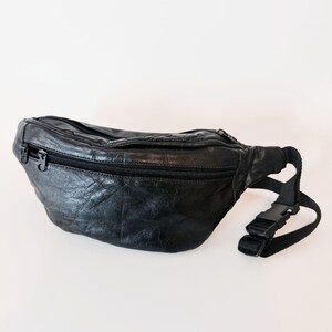 May include: A black leather fanny pack featuring a textured surface, a horizontal zippered main compartment with two black metal zipper pulls, and an adjustable black nylon webbing strap secured with a black plastic side-release buckle closure.
