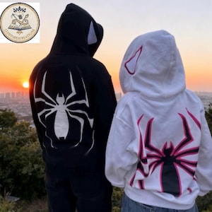 May include: A black hoodie featuring a large white spider graphic on the back and a white hoodie featuring a large black and pink spider graphic on the back. Both hoodies have hoods with small ear-like accents, shown from the back view.
