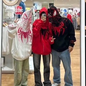 Peut inclure: Trois sweats à capuche zippés avec des motifs inspirés de Spider-Man. L'un est blanc avec des motifs de toile d'araignée roses, un est rouge uni avec un emblème d'araignée noir, et un est noir avec des toiles rouges et un grand motif d'araignée.