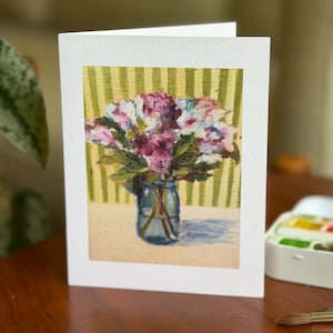 May include: A white greeting card featuring a printed painting of a bouquet of pink and white flowers in a blue glass jar. The background of the painting shows vertical green and white stripes, and the surface is a light beige color.