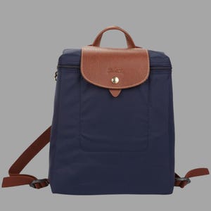 May include: A navy blue backpack featuring a brown leather flap with a gold-tone snap button closure. The bag includes a top handle, adjustable brown shoulder straps, and a zipper closure along the top edge, constructed from a smooth fabric material.