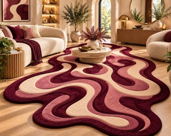 Op de afbeelding: Een vloerkleed met een onregelmatige, organische vorm en gelaagde, golvende banen in tinten diep bordeaux, gedempt roze en crème. Het kleed heeft een dikke, getextureerde pool en ligt op een lichte houten vloer in een woonkamer.