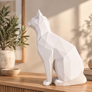 May include: A white cat figurine featuring a geometric, faceted design. The sculpture is rendered in a solid matte white finish with sharp, angular planes that define the feline form, including pointed ears, a sleek neck, and a seated posture.