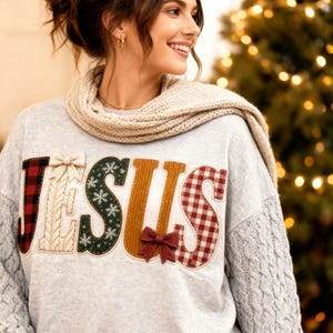 May include: A light gray long-sleeve sweatshirt featuring the word JESUS in large, textured applique letters. Each letter has a unique pattern, including red and black buffalo plaid, cream cable knit, green with white snowflakes, and red gingham.