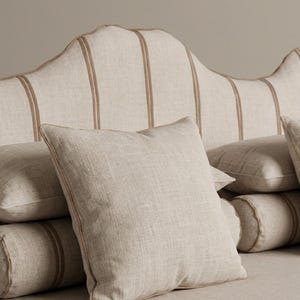 Può includere: Una testiera imbottita color beige con bordo superiore smerlato e profili verticali in una tonalità marrone più scura. Diversi cuscini quadrati e cilindrici nello stesso tessuto beige con profili abbinati sono disposti davanti alla testiera.