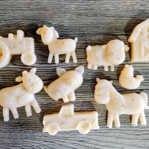 May include: A collection of nine off-white, textured soap bars shaped like farm animals and vehicles, including a tractor, goat, sheep, barn, cow, pig, chicken, horse, and pickup truck, all featuring small embedded botanical particles throughout.