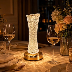 May include: A cylindrical table lamp with a clear, diamond-cut textured glass shade that emits a warm, glowing light. The lamp features a circular gold-toned metal base and sits on a dining table alongside two wine glasses and a floral arrangement.