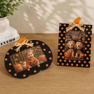 Può includere: Due cornici nere a pois arancioni. Una è a forma di zucca con un fiocco in rafia e la foto di un bambino. L'altra è rettangolare con una decorazione a zucca, un fiocco in rafia e la foto di due cuccioli con cappelli da strega.