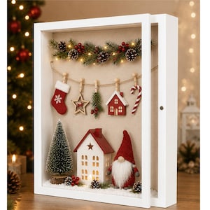 May include: A white rectangular shadow box frame containing a Christmas display. Inside, a string holds a red stocking, a wooden star, a pine sprig, a red house, and a candy cane. Below, a miniature pine tree, a white house, and a gnome stand.