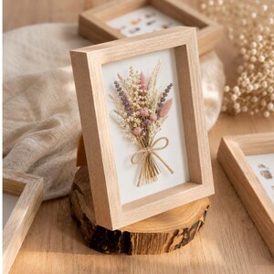 Peut inclure: Un cadre rectangulaire en bois clair contenant un bouquet pressé de fleurs séchées dans des tons rose, lavande et crème. L'arrangement est noué avec un fin ruban de jute et placé sur un fond blanc à l'intérieur du cadre vitré.