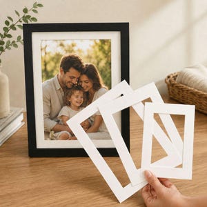 May include: A black rectangular picture frame containing a photograph of a family of three, accompanied by three white cardboard mat boards of varying sizes held in a hand, designed for framing and displaying photographic prints.
