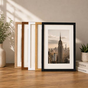 May include: A set of five rectangular picture frames arranged in a row, featuring various finishes including light wood, dark wood, white, natural oak, and black. Each frame contains a white mat board and a photograph of the Empire State Building.