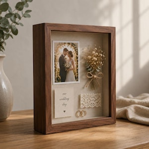 May include: A rectangular wooden shadow box frame containing a photograph of a couple, a small dried flower bouquet tied with twine, a piece of white lace, two gold rings, and a card with the printed text our wedding day and a small heart.