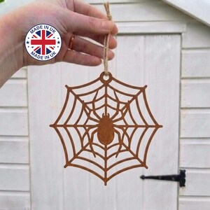 May include: A brown, laser-cut wooden ornament featuring a spider sitting in the centre of a web. The ornament is suspended by a thin, light brown twine loop. A circular sticker with the text "MADE IN UK" is visible in the upper left corner.