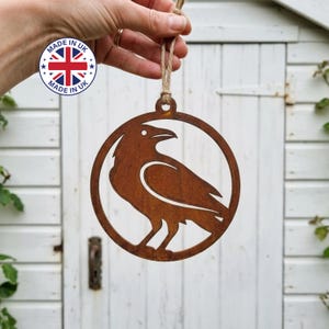 May include: A circular metal ornament featuring a cutout silhouette of a raven. The piece has a weathered, rusted brown finish and includes a small loop at the top attached to a piece of natural jute twine for hanging purposes.