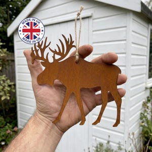 May include: A flat, rust-coloured metal moose silhouette ornament featuring a textured, weathered finish. The moose is suspended by a thin, light brown jute twine loop attached to the top of its back, designed for hanging as a decorative piece.