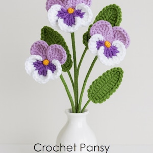 May include: Three crocheted pansy flowers with purple, white, and yellow centres on green stems with green leaves, arranged in a small white vase. The text Crochet Pansy PDF PATTERN is printed in black below the flowers.