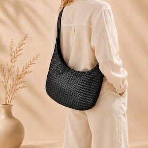 May include: A black shoulder bag featuring a woven leather texture and a curved silhouette. The bag is worn over the shoulder with a wide strap, showcasing a consistent grid-like pattern across the entire exterior surface of the accessory.