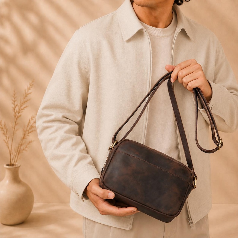 May include: A dark brown leather crossbody bag with a rectangular shape, a smooth matte finish, and a thin adjustable shoulder strap. The bag features visible stitching along the edges and metallic hardware connecting the strap to the main body.