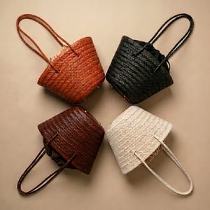 May include: Four woven bucket bags arranged in a square formation. Each bag features a distinct color: tan, black, dark brown, and cream. The bags have braided handles and a visible interior lining, showcasing a consistent woven texture throughout.