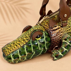 May include: A handbag shaped like a crocodile with a textured green and yellow scale pattern. The bag features brown leather handles, a gold-tone metal zipper closure, and a dark brown eye detail with a yellow pupil on the side of the snout.