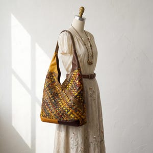 Puede incluir: Un bolso de hombro estilo hobo con un patrón de cuero trenzado multicolor en tonos amarillo, marrón, azul y rojo. El bolso incluye paneles de cuero sólido en marrón y amarillo, una correa de hombro única y una silueta amplia y holgada.