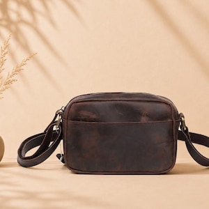 May include: A dark brown leather crossbody bag with a rectangular shape, a top zipper closure, and an adjustable shoulder strap attached with antique brass-toned metal hardware. The bag features a smooth, matte finish with subtle natural variations.