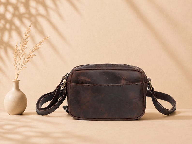 May include: A dark brown leather crossbody bag with a rectangular shape, a top zipper closure, and an adjustable shoulder strap attached with antique brass-toned metal hardware. The bag features a smooth, matte finish with subtle natural variations.