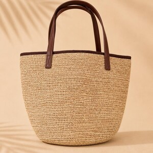 Pu&ograve; includere: Una borsa tote strutturata con corpo in paglia intrecciata color marrone chiaro e base arrotondata. La borsa presenta manici in similpelle marrone scuro e un bordo coordinato lungo il profilo superiore, creando un contrasto con la trama naturale.