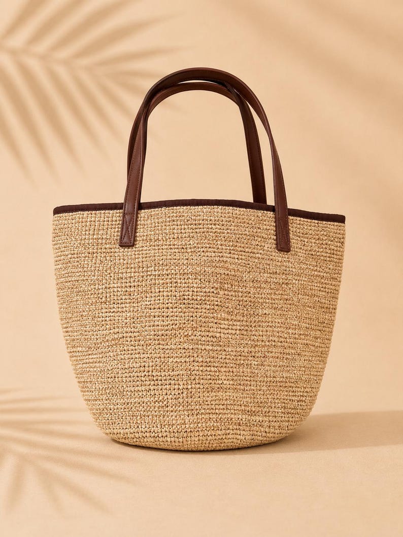 Pu&ograve; includere: Una borsa tote strutturata con corpo in paglia intrecciata color marrone chiaro e base arrotondata. La borsa presenta manici in similpelle marrone scuro e un bordo coordinato lungo il profilo superiore, creando un contrasto con la trama naturale.