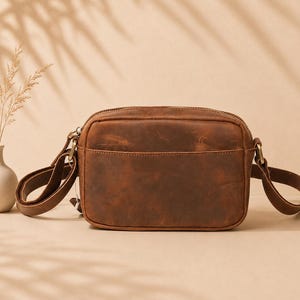 May include: A rectangular brown leather crossbody bag with a smooth, matte finish and a visible top zipper closure. The bag features a front slip pocket, matching leather side straps, and antique brass-toned metal hardware attachments.