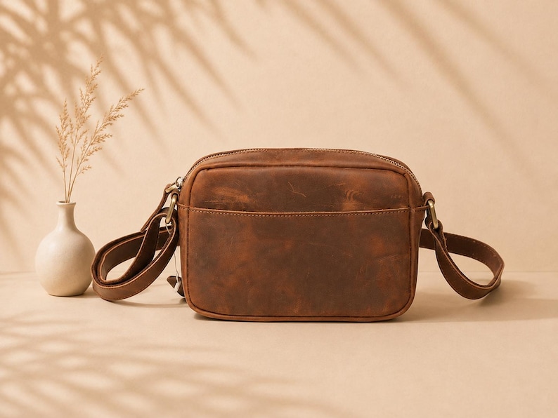 May include: A rectangular brown leather crossbody bag with a smooth, matte finish and a visible top zipper closure. The bag features a front slip pocket, matching leather side straps, and antique brass-toned metal hardware attachments.