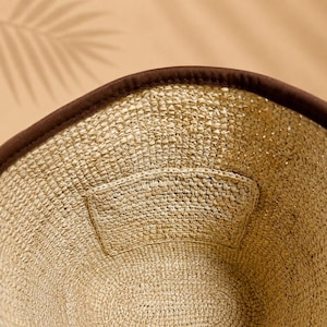 Pu&ograve; includere: Un cappello da pescatore in paglia intrecciata color beige naturale con un bordo in tessuto marrone scuro. L'interno del cappello presenta una toppa in paglia intrecciata coordinata cucita sul lato, che mette in risalto la trama del tessuto.